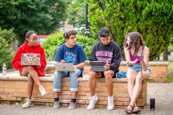 Four students sitting on a low brick wall on a sunny green quad. One girl is holding up her phone and gesturing toward it while the other three, using their laptops, look over at what she's showing them.