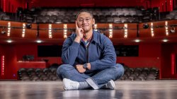 Josh De La Cruz sitting cross-legged on a stage in a proscenium theater