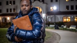 Horatio Wauchope , wearing a winter coat, stands outside on a campus walkway at night, holding a large folder to his chest in front of a lit academic building.