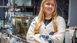 Kelly Witters stands in a marine biology lab with arms crossed, in front of stacked aquariums and tubing used for aquatic research.