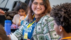 Isabella Zarate Gonzalez, wearing a blue SPARK T-shirt and a green-and-white crochet cardigan, sits at a table holding a worksheet, speaking with an elementary student who is seated around her during a classroom activity.