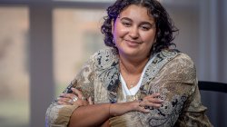 Dani Mazariegos sits at a studio desk with her arms folded, wearing a patterned blouse.