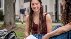 Dana Brzezinski sits on the grass at Montclair State University, holding an open notebook and talking with a friend, with campus buildings and a tree visible in the background.
