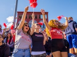 Three female students in a large crowd at homecoming cheering for the Montclair football team