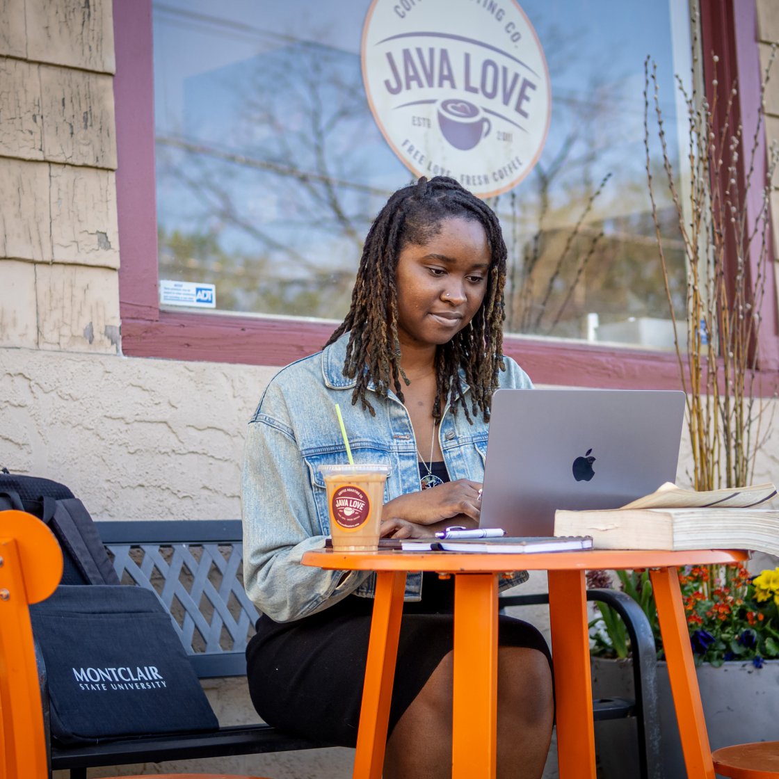 Female adult student sitting at a table outside cafe, with coffee and a laptop in front of her.