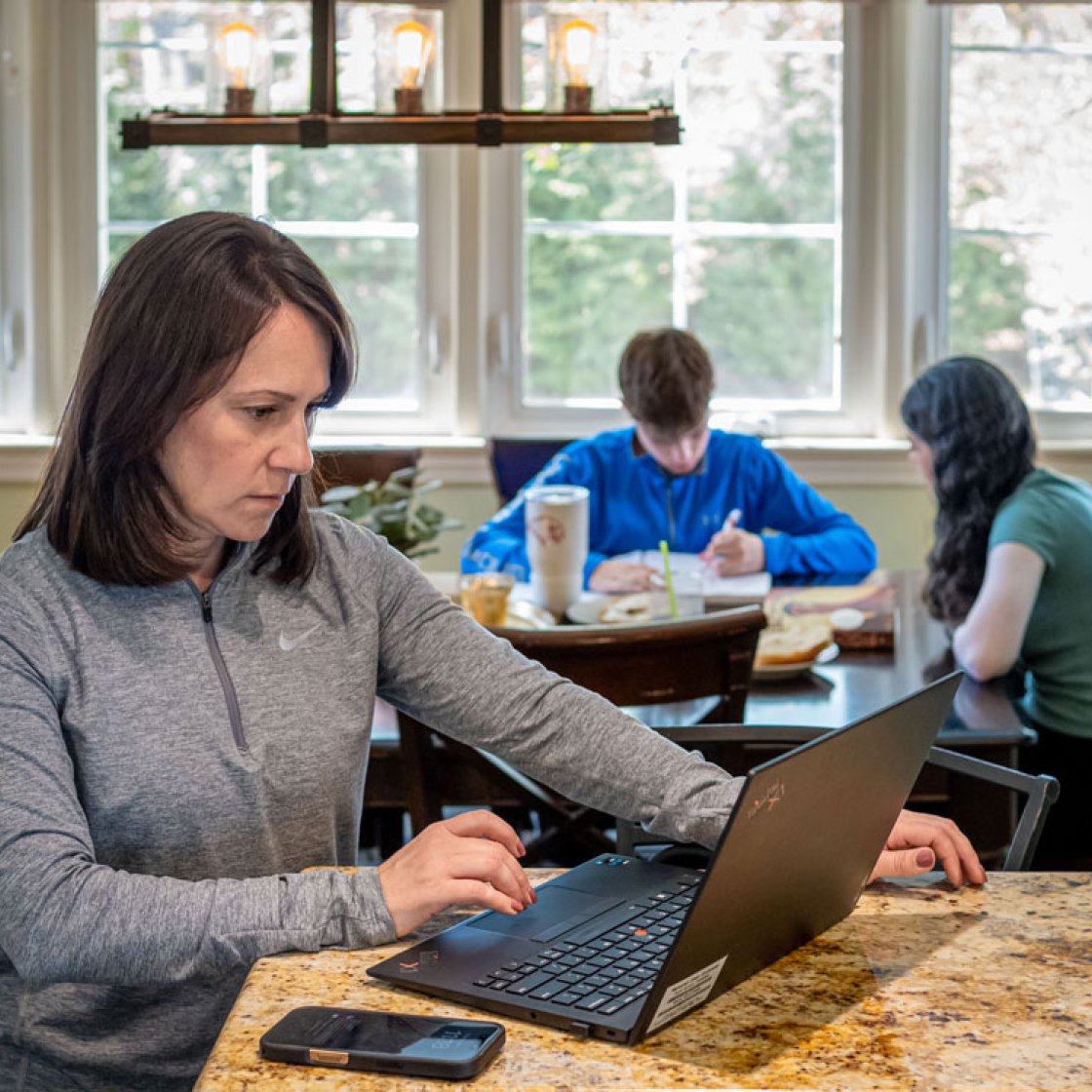 Adult Montclair Online student works on a laptop at a kitchen counter while her two teenagers do homework at the dining table in the background.