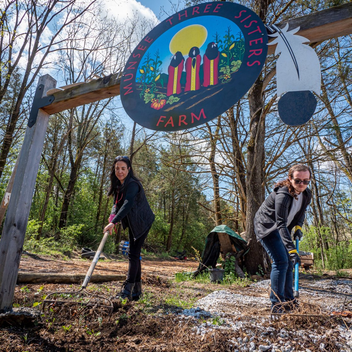 Two people are working with gardening tools beneath a large entry sign at Munsee Three Sisters Farm.