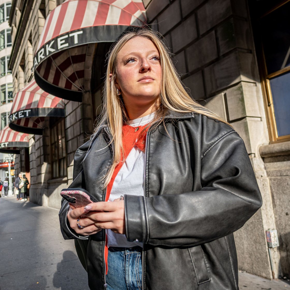 Emma Haskell stands outside a New York City subway stop, holding her phone, with a cityscape visible in the background.
