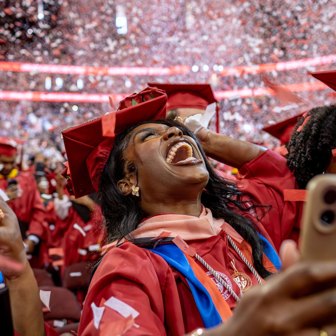 A female student in a red cap and gown joyfully smiles while looking up at confetti falling down at commencement. 