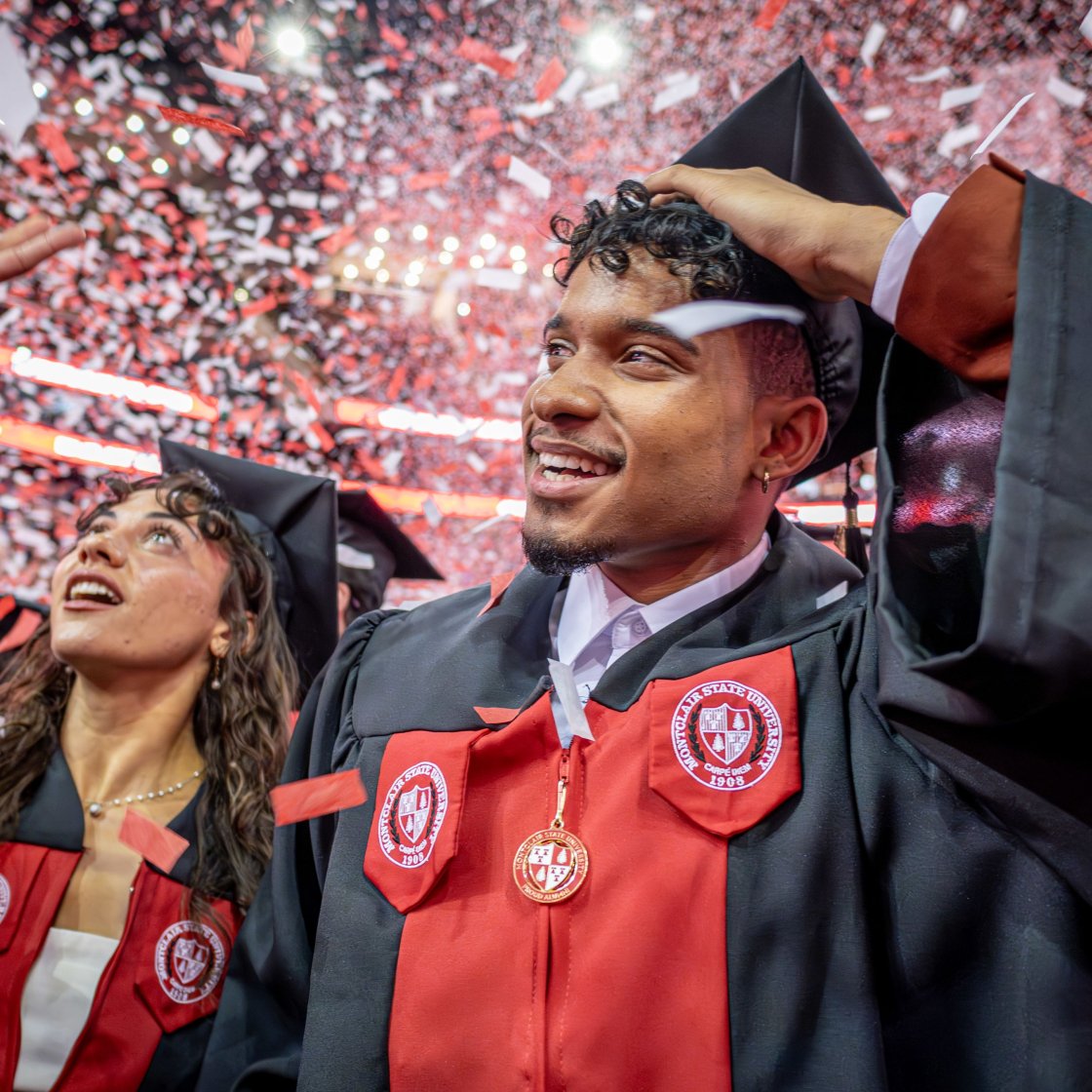 Two students, a female and male, in black caps and gowns smile as confetti falls during commencement. 
