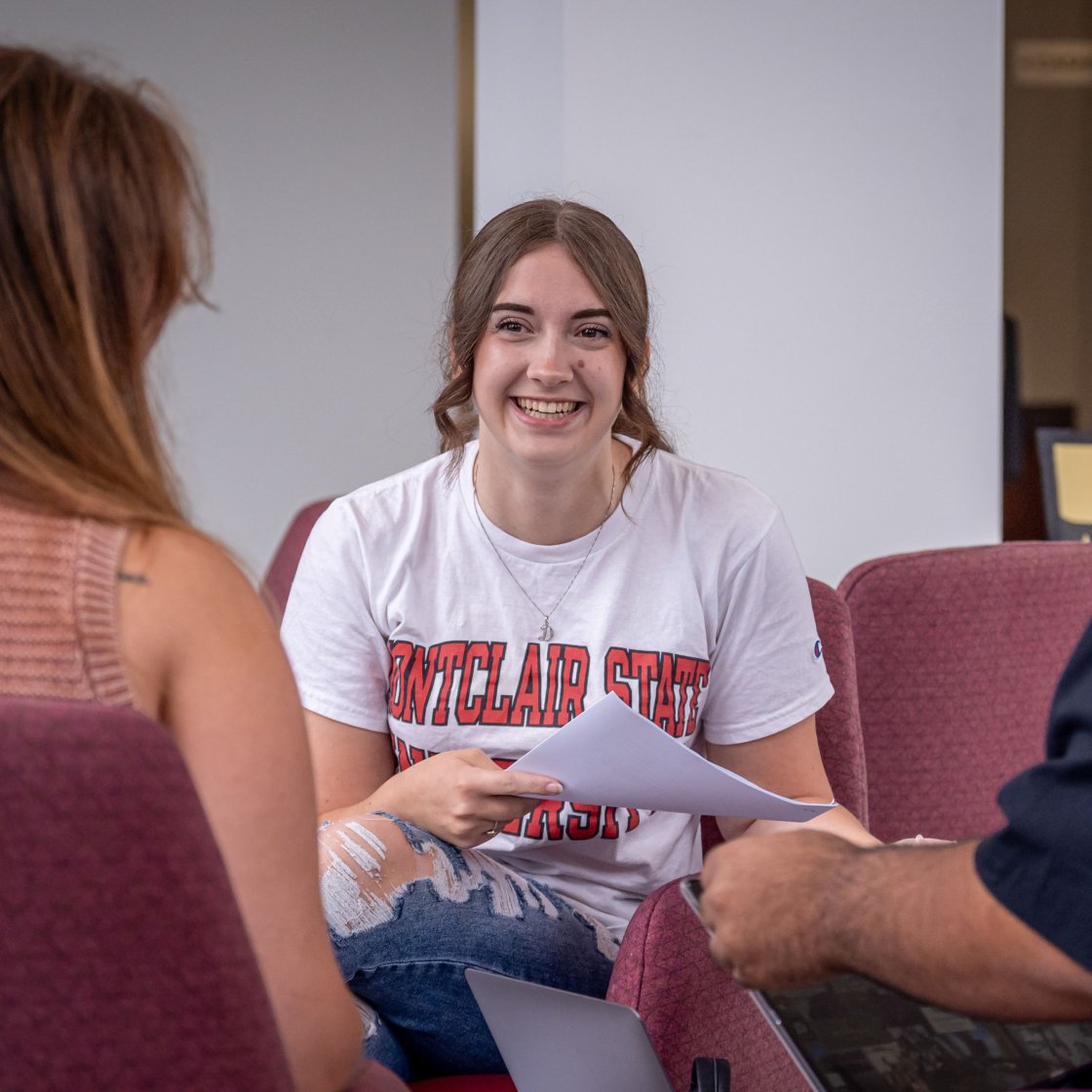 Three students enjoying a conversation; one in the center wears a Montclair sweatshirt and the student on the right is laughing.
