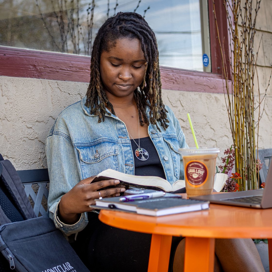 Online student sitting outside cafe, reading a book with an open laptop on the table in front of her.