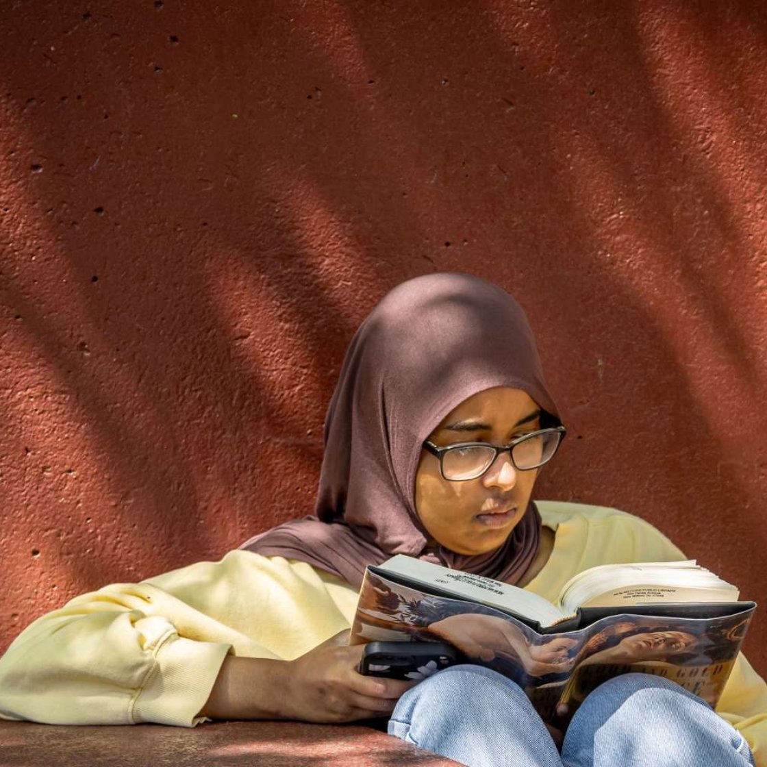 Student wearing a hijab and reading a book in the shade.