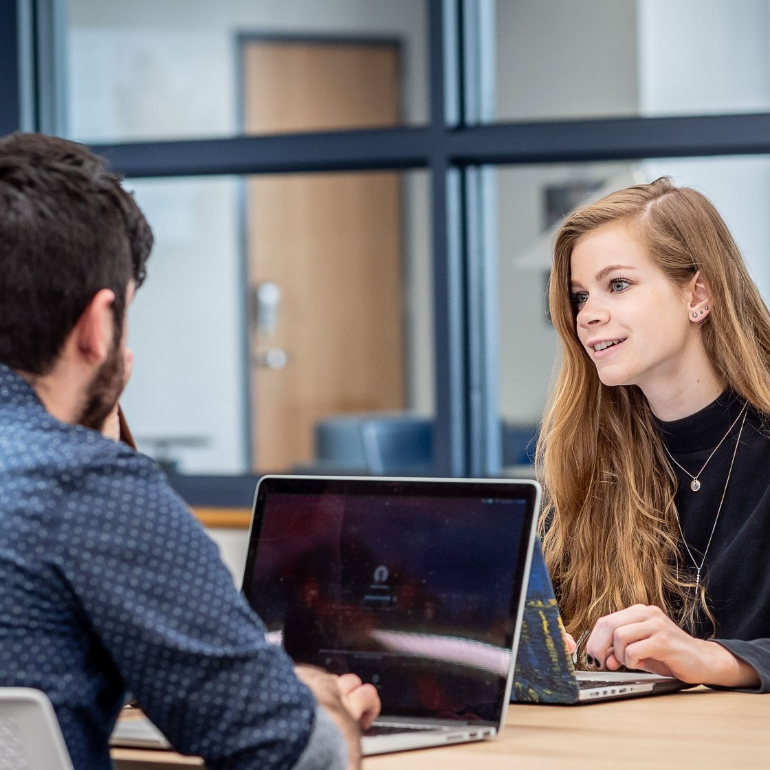 Two students sit across from each other with laptops open on the table in between them.