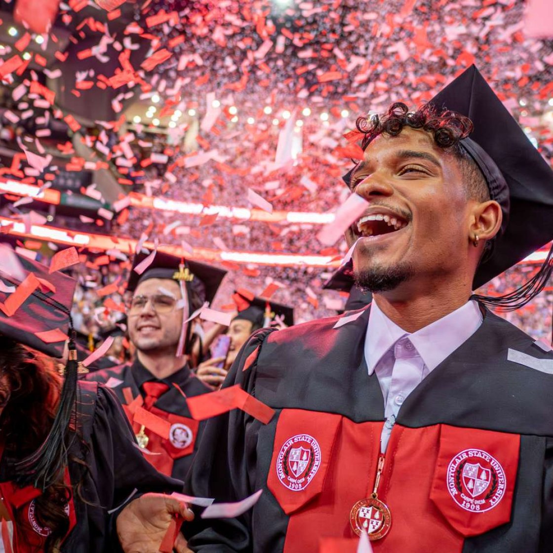 students smiling at graduation ceremony wearing caps and gowns as confetti falls.