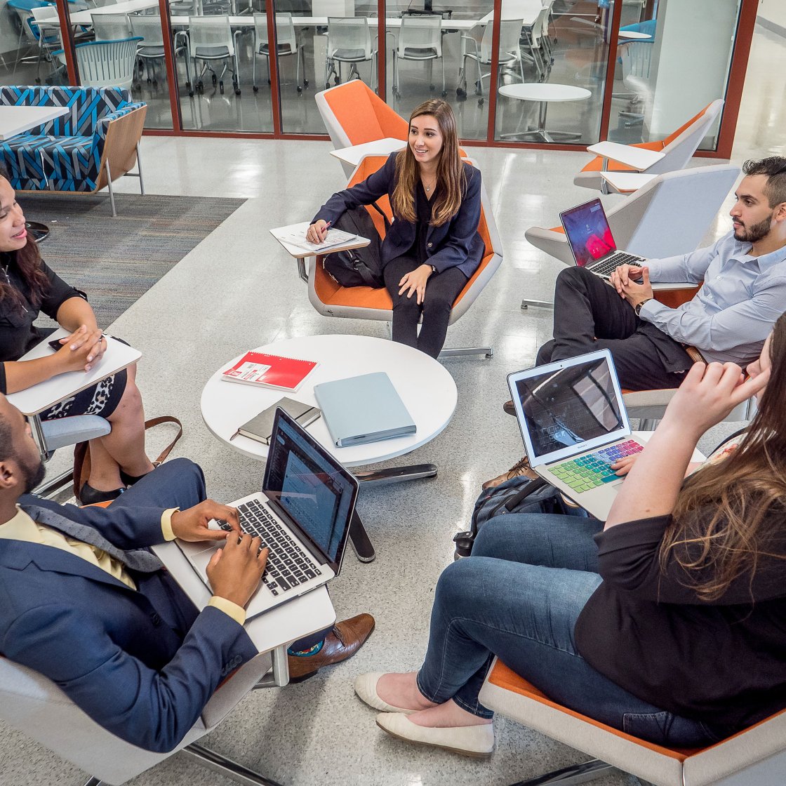 Five people are seated in a modern lounge area, around a small white table. Each person sits in an orange chair and is engaged with laptops, notebooks, or paper folders.