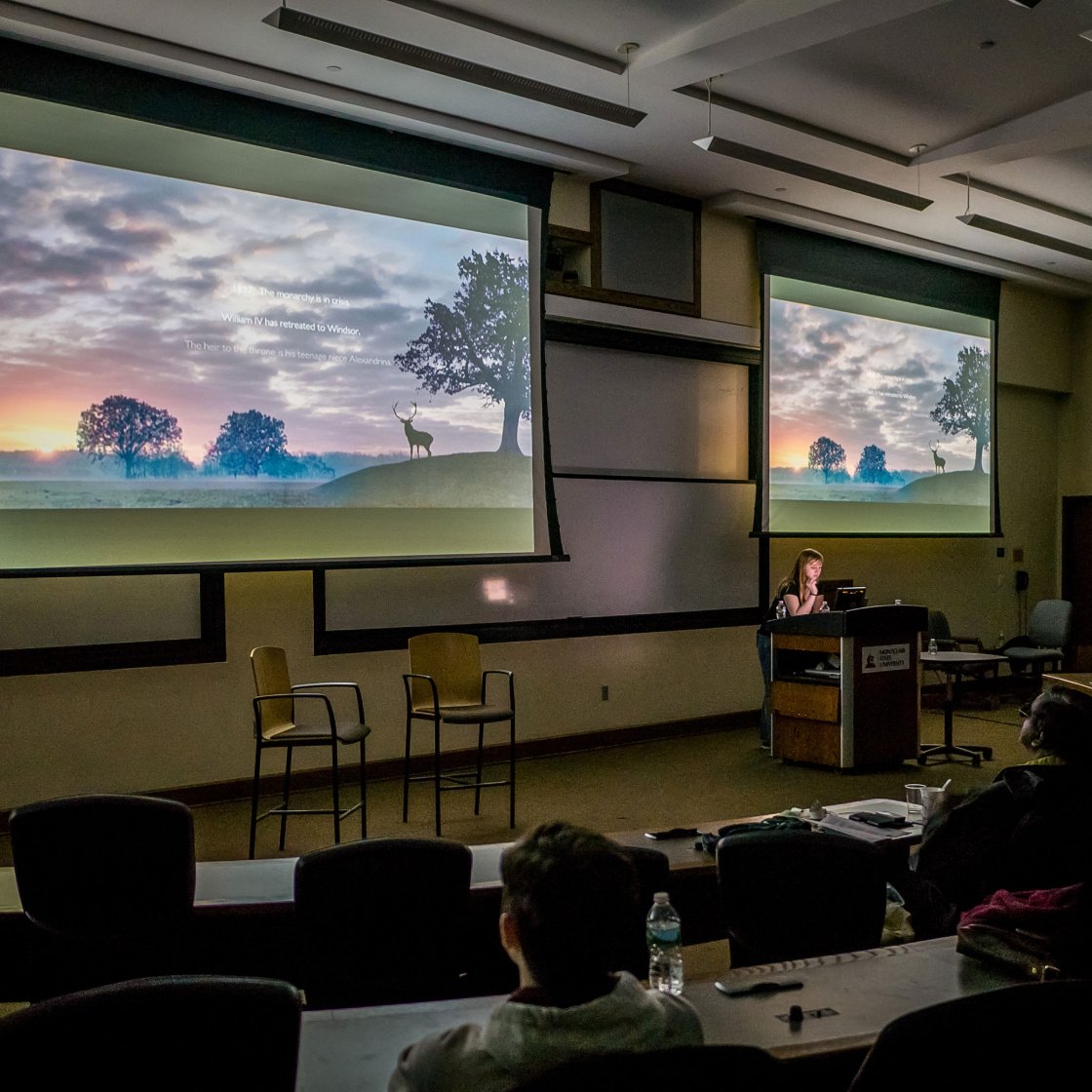 Students watch a film on projector screens in a darkened lecture hall.