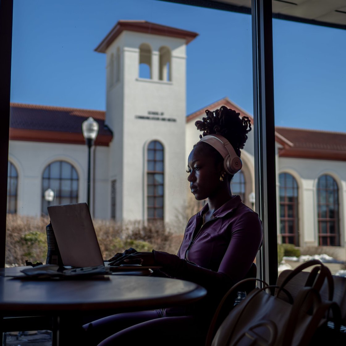 A student works on a laptop in front of a large window. An academic building is in the background.