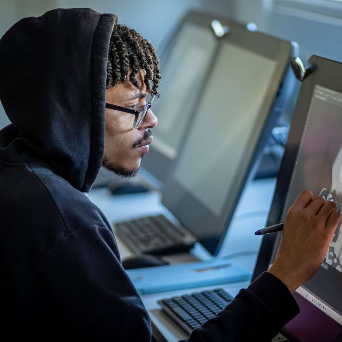 Illustration student wearing a hoodie and glasses drawing on a digital screen.