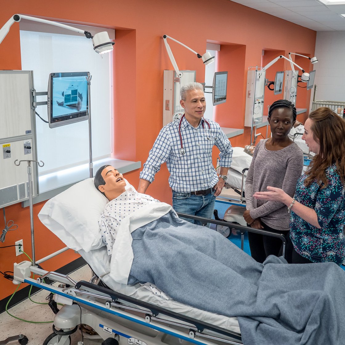 Three students talk at the bedside of a mannequin for realistic medical simulations.