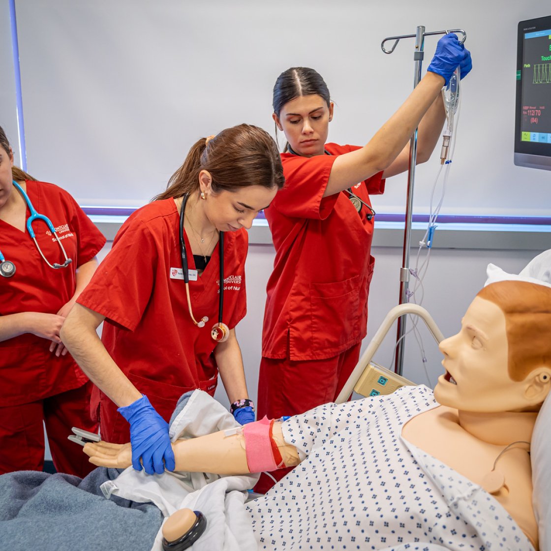 Three nursing students in red scrubs practice clinical skills on a hospital mannequin, adjusting an IV line and monitoring vital signs on a screen.