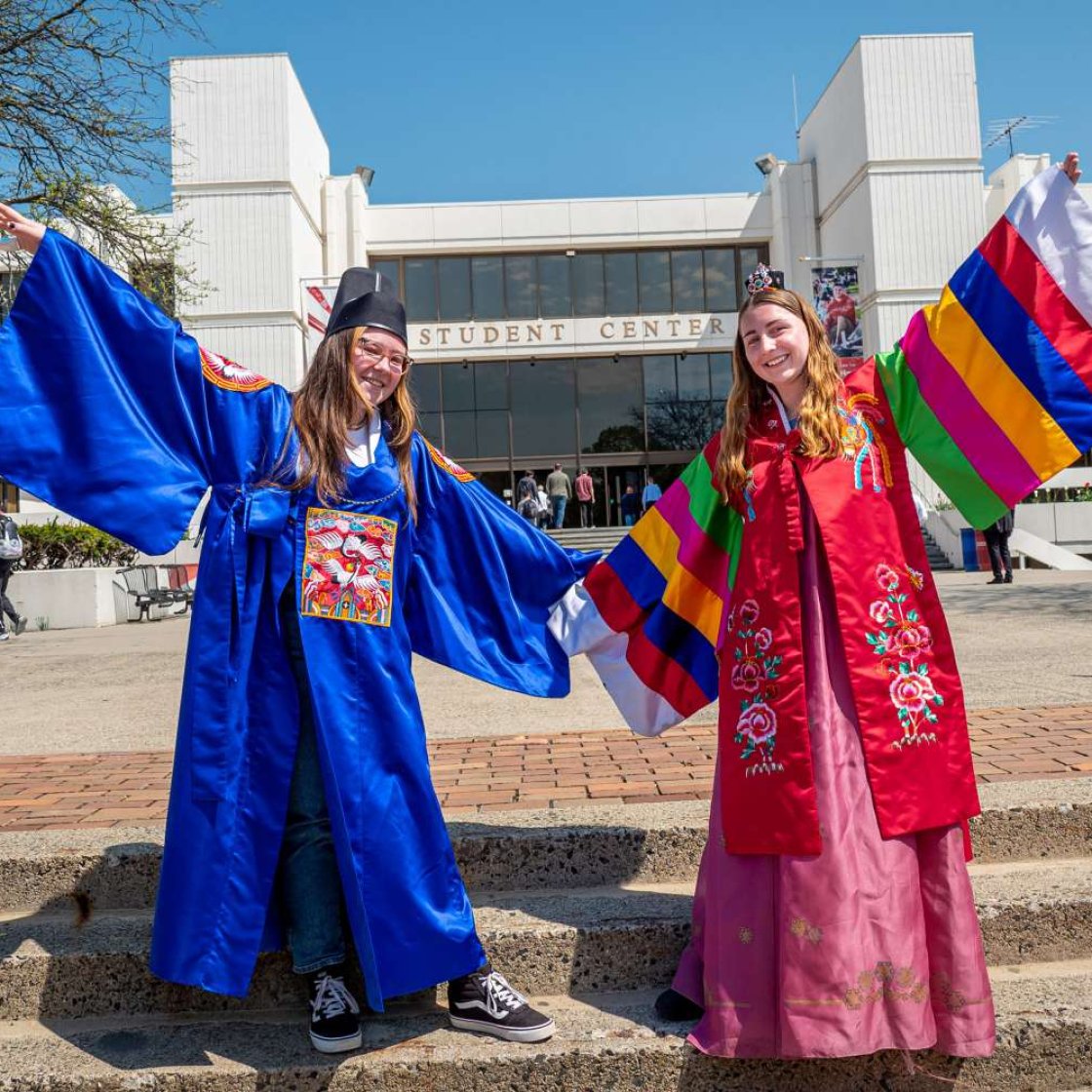 Two students wearing colorful traditional Korean hanbok and robes posing with arms outstretched in front of the Student Center.