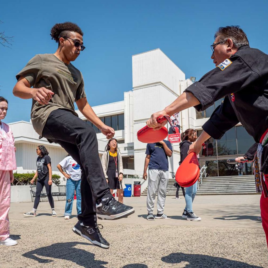 Student practices a high kick during a taekwondo demonstration outdoors at a campus event. 