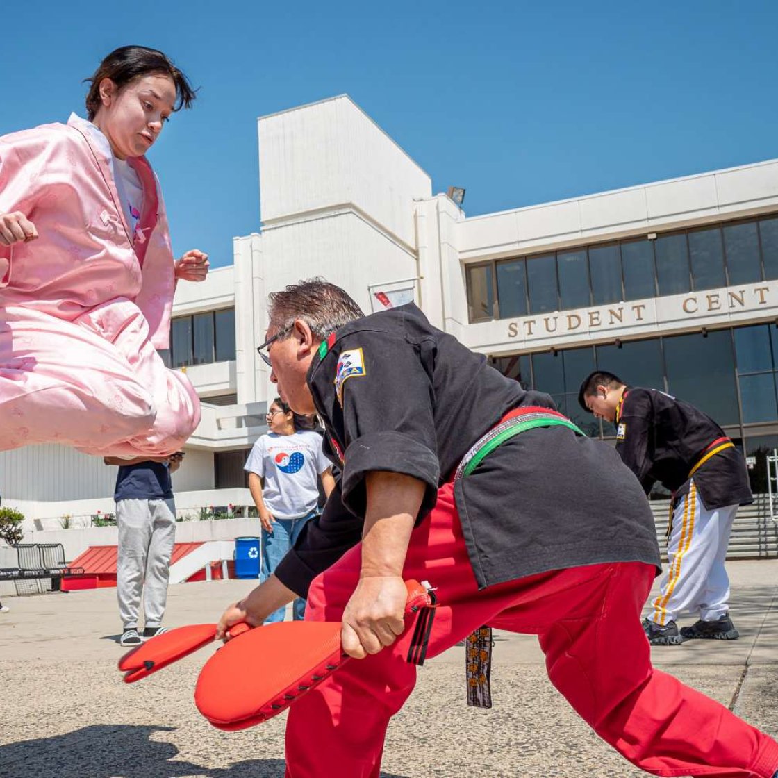 A student dressed in a pink martial arts uniform performs a high jump kick during a Taekwondo workshop held outdoors in front of the Student Center, while an instructor in a black uniform holds a target pad.