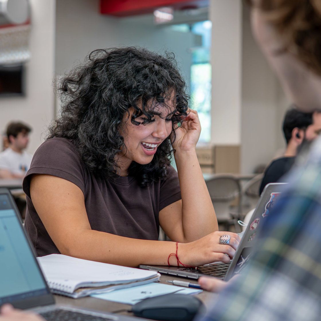 A student laughs while working on a laptop with other students in a classroom.