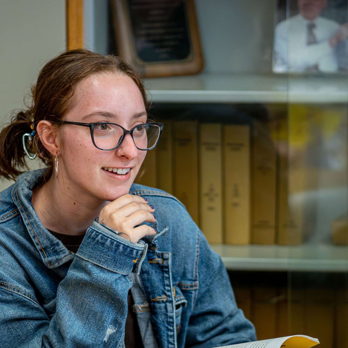 A smiling student sits in a classroom with an open book on the table in front of them.