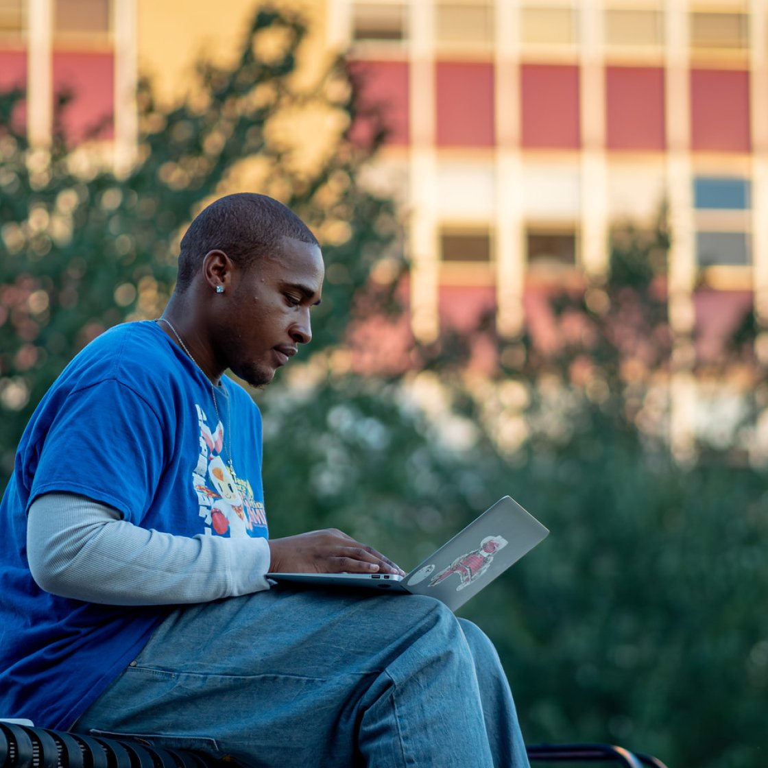 Student in a blue shirt sits outdoors on campus, focused on working on a laptop with a Red Hawk sticker.