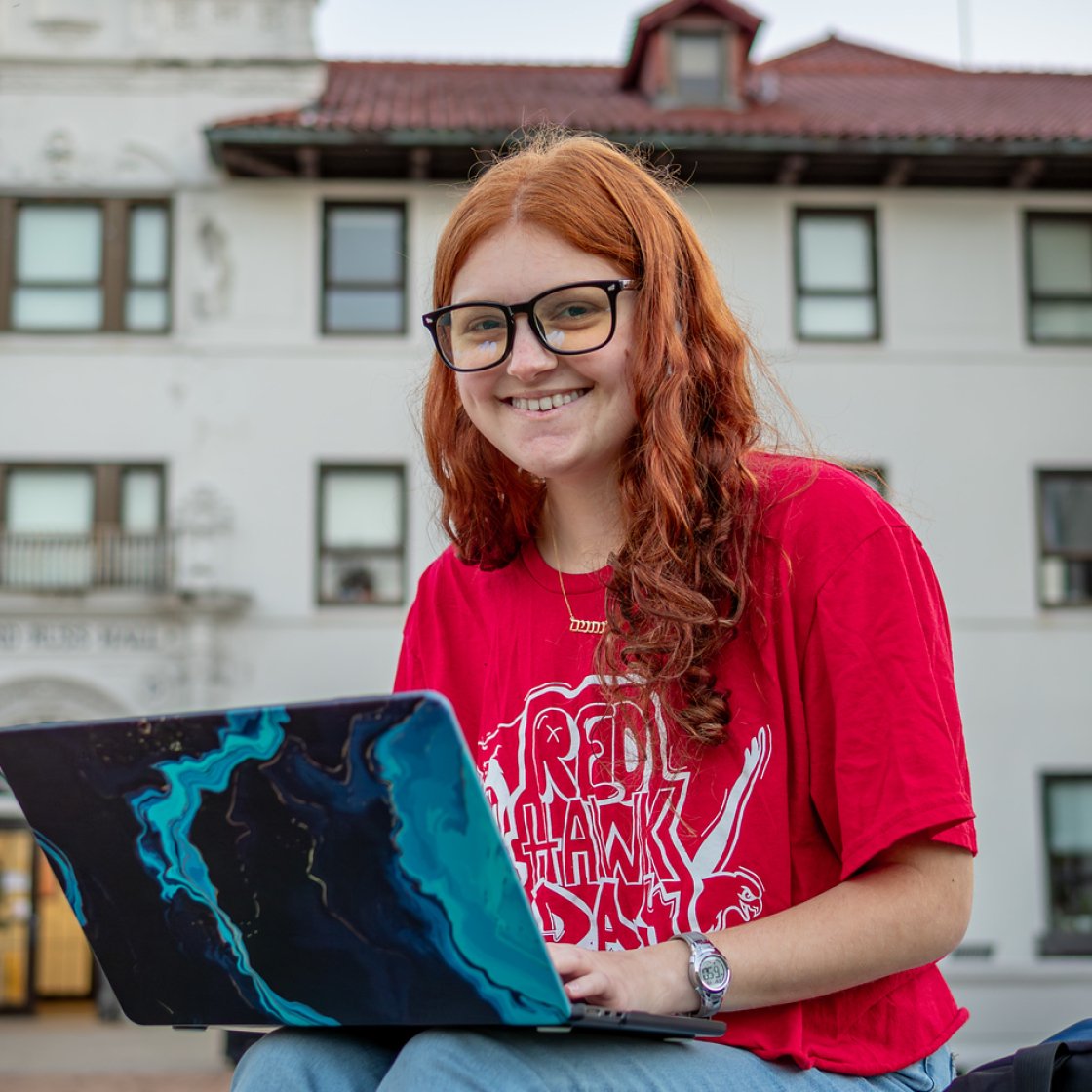 Student wearing a Montclair Red Hawk t-shirt smiles while working on a laptop outside an academic building during summer.
