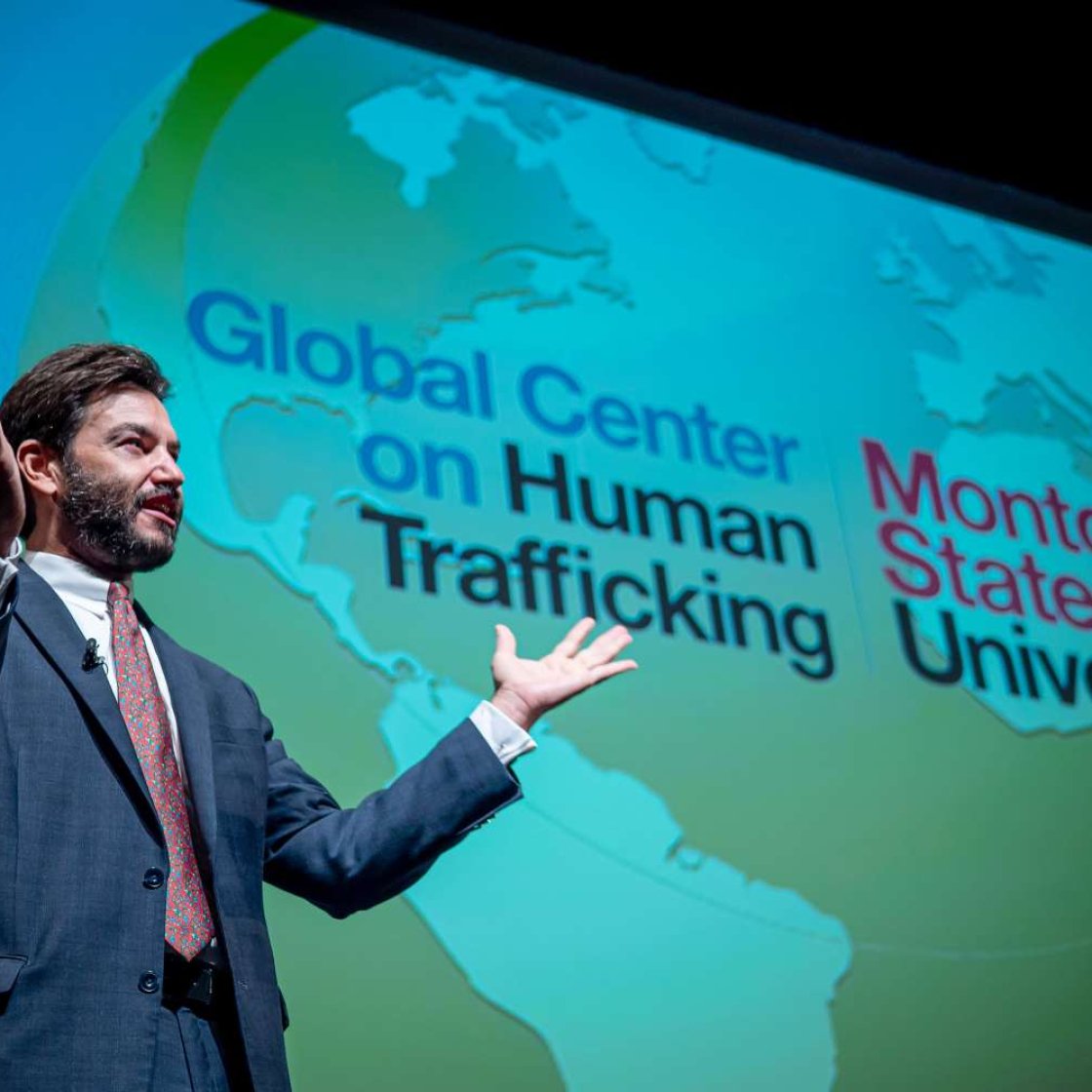 Montclair State University President Jonathan Koppell stands on stage in front of a large screen displaying a world map and the words "Global Center on Human Trafficking" and "Montclair State University."