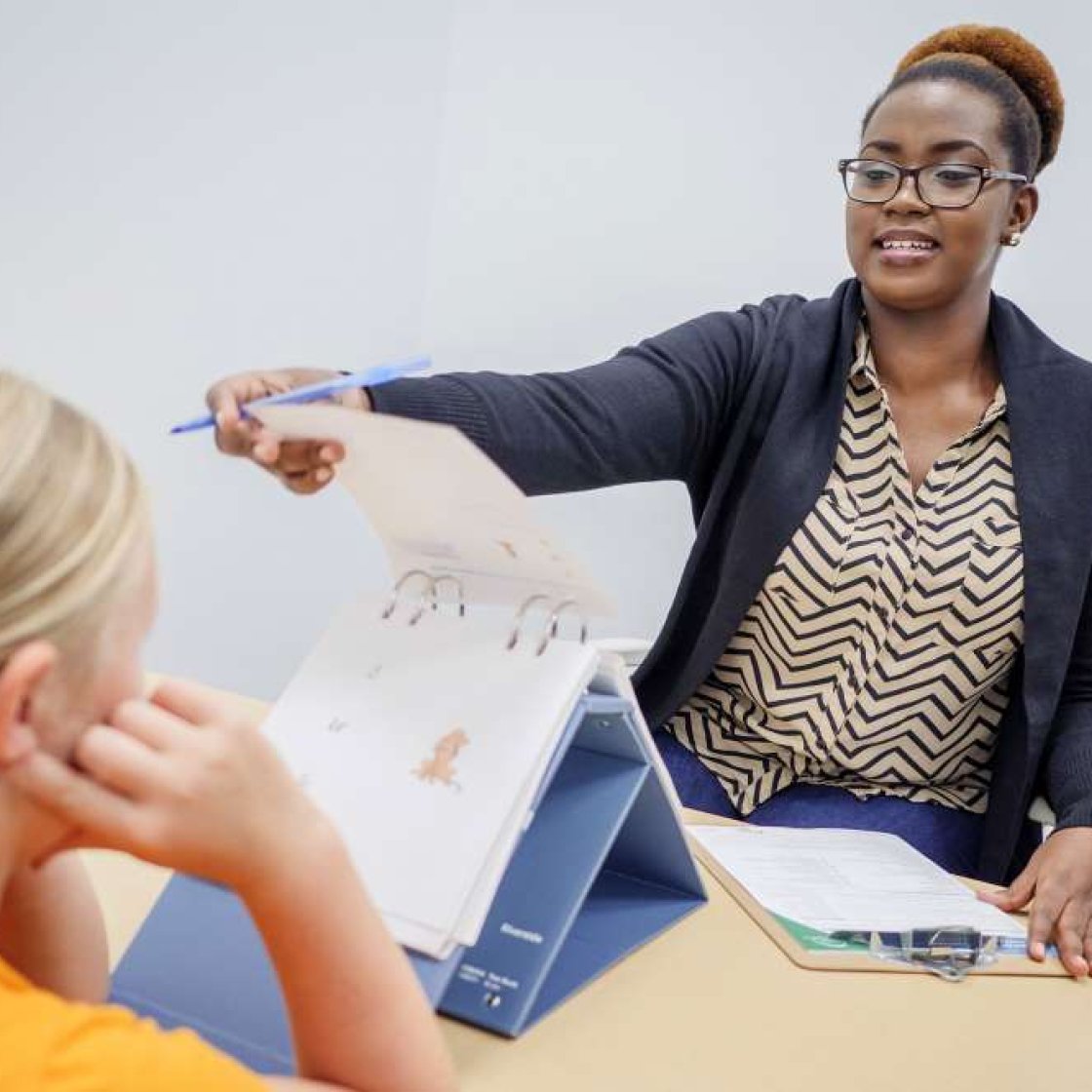 A clinician holds up a page of illustrated images and points to it while assessing a child seated across the table. Assessment materials and notes are visible on the table in a professional clinical setting.