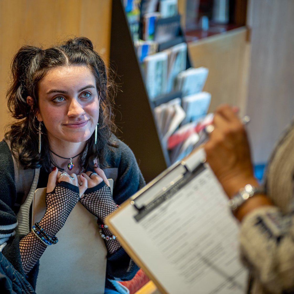 A student with dark hair and a backpack stands in a hallway, speaking to someone holding a clipboard with papers; bookshelves are visible in the background. 