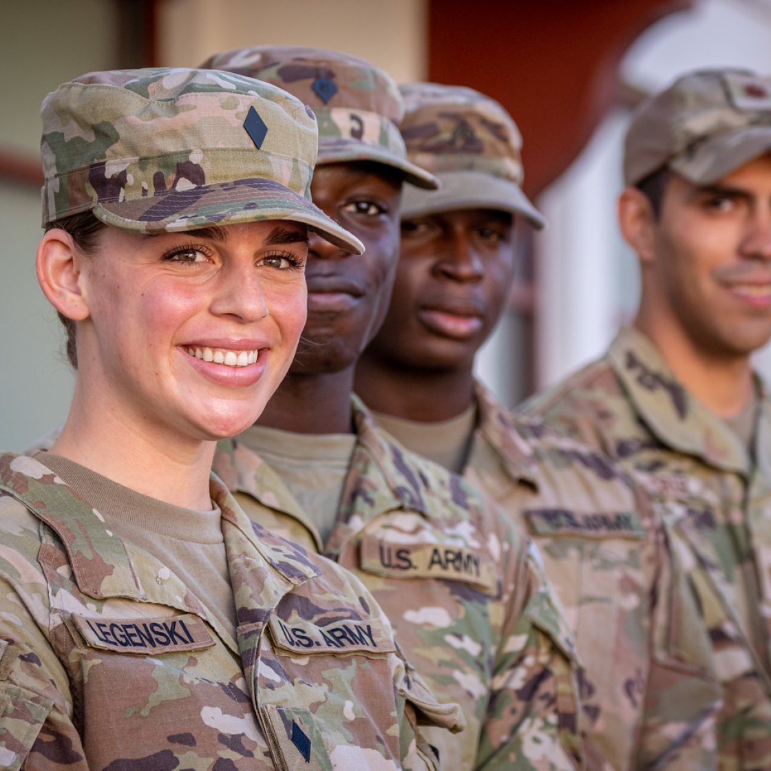 Several U.S. Army soldiers in camouflage uniforms and caps stand in formation outdoors. 