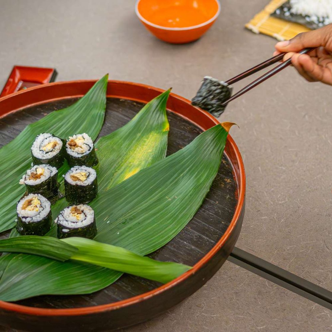 A close-up of a person using chopsticks to pick up a sushi roll from a round tray lined with large green Aspidistra leaves, with several pieces of sushi arranged on the tray. In the background, a bamboo sushi rolling mat with rice and an orange bowl are visible on the table.