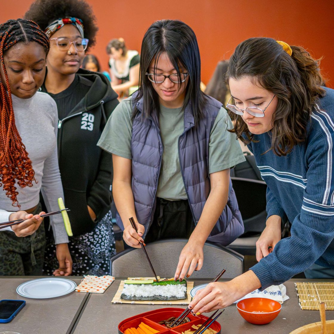A group of students stands around a table making sushi using bamboo mats and hands, picking up fillings with chopsticks. Various sushi ingredients such as cucumber and carrot sticks, egg slices as well as bowls and plates are arranged on the table. The activity takes place in a classroom with others participating in the background.