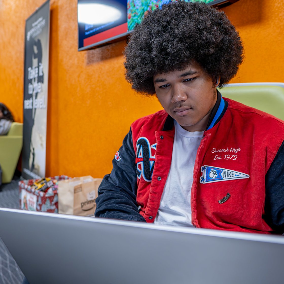 A student wearing a red and black varsity jacket works on a laptop in a campus lounge with bright orange walls. 