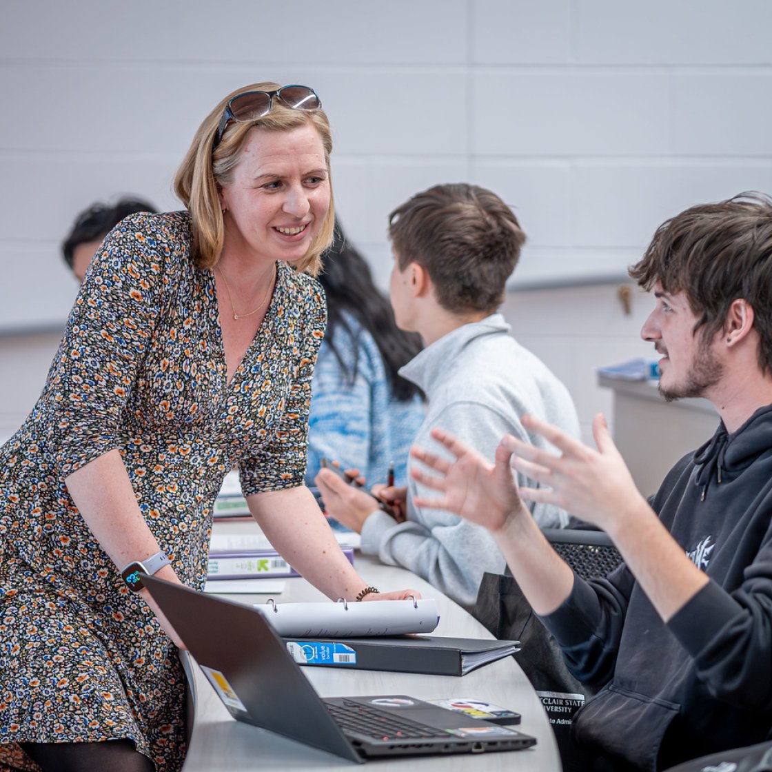 Instructor leans over a desk to talk with a student in a classroom, as other students sit nearby and a laptop and notebooks are spread out on the table.