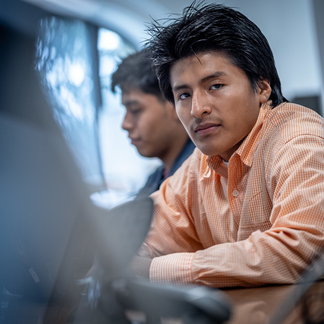 Student wearing an orange button-down shirt leans forward while working on a computer screen.