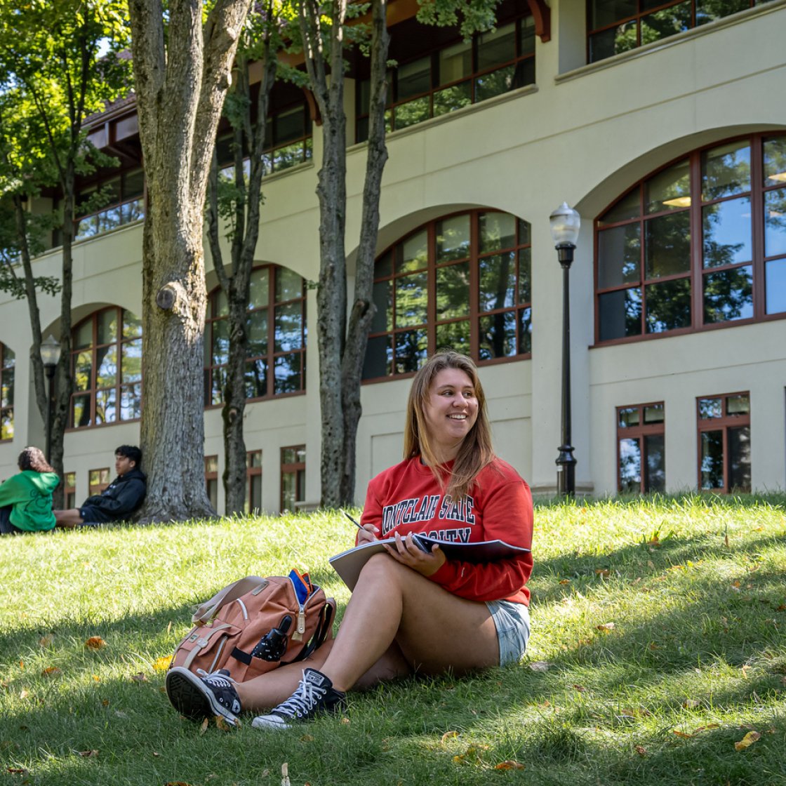 Student sitting on a grassy lawn in front of a large campus building with arched windows, wearing a red university shirt and writing in a notebook.