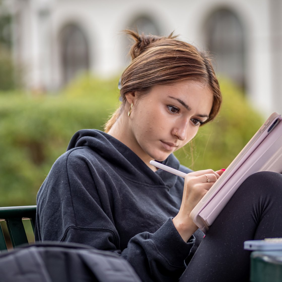 Student sitting outdoors on a campus bench, wearing a dark hoodie and leggings, focused on writing in a notebook or textbook balanced on their knees.