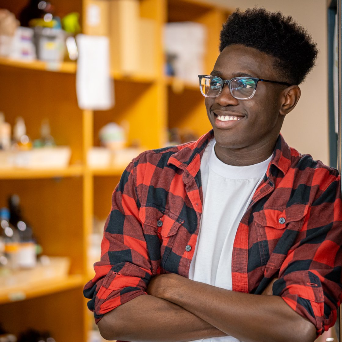 Student standing with folded arms in a doorway, wearing a red-and-black plaid shirt over a white T-shirt, with shelves softly blurred in the background.
