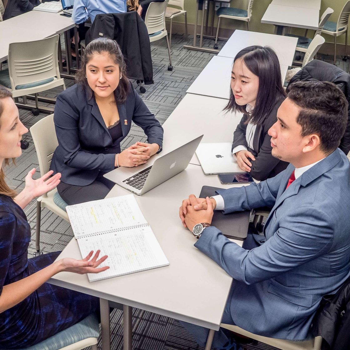 Four students sit at a table with notebooks and laptops open between them.