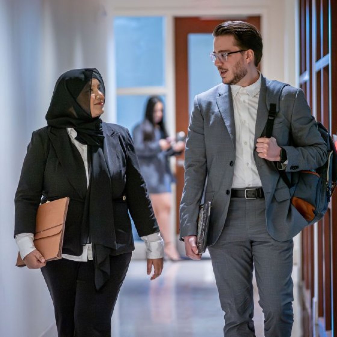 Two students in business attire, walking and talking in a hallway.
