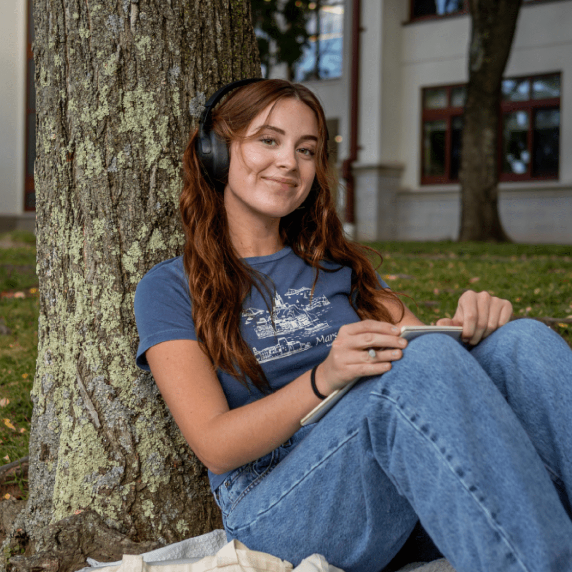 Student sitting down against a tree with headphones on smiling.