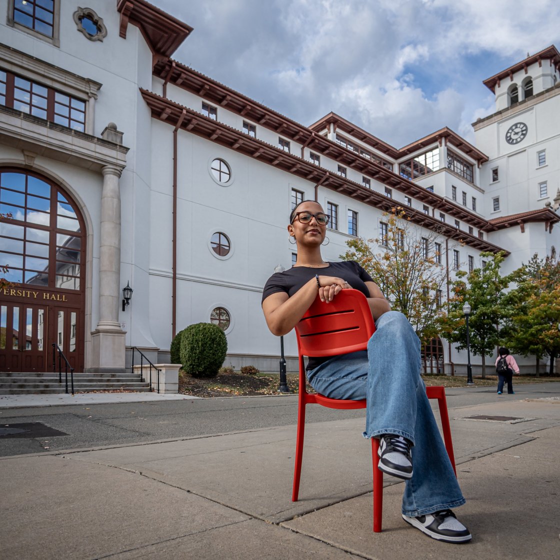 A student sits in a red chair in front of an academic building.