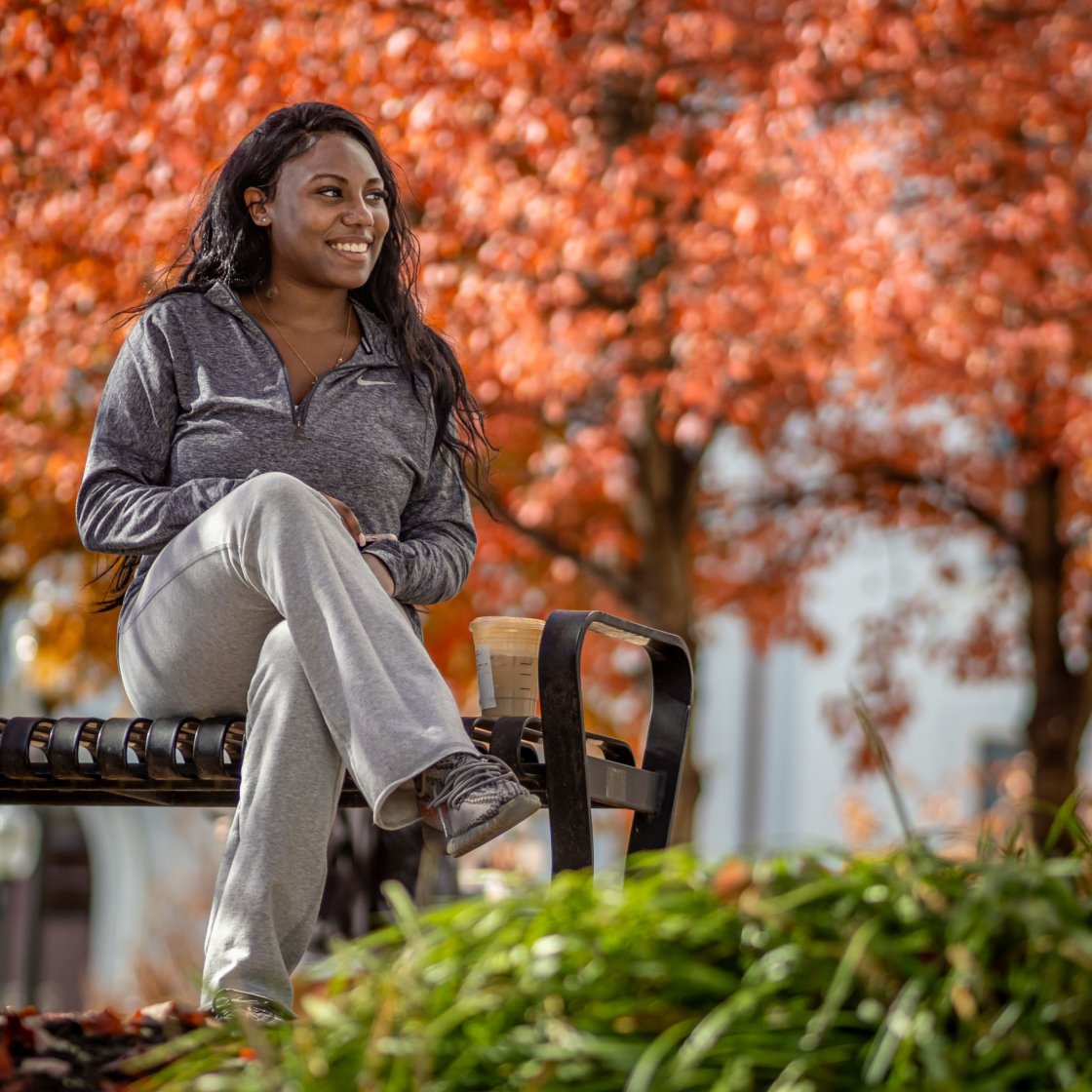 Female student is sat on a bench surrounded by trees with orange leaves.