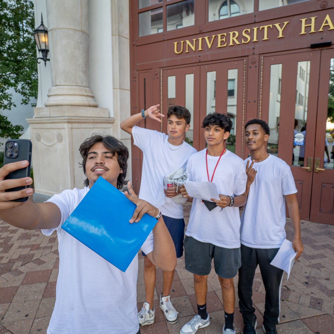 Four students are seen posing in front of University Hall taking a picture.